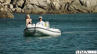 Kendra Sunderland takes standing doggy on a rocky shore with blue water behind her